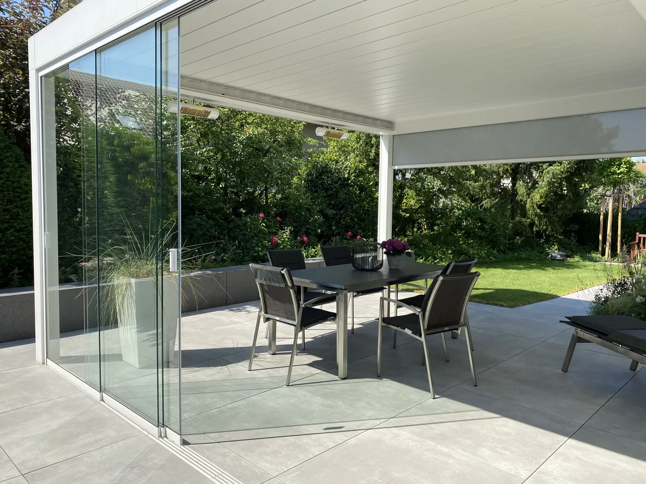 Moderne Terrassenüberdachung mit Glas-& Schiebeelementen, großformatigen Terrassenplatten und fließendem Übergang zwischen Haus und Garten